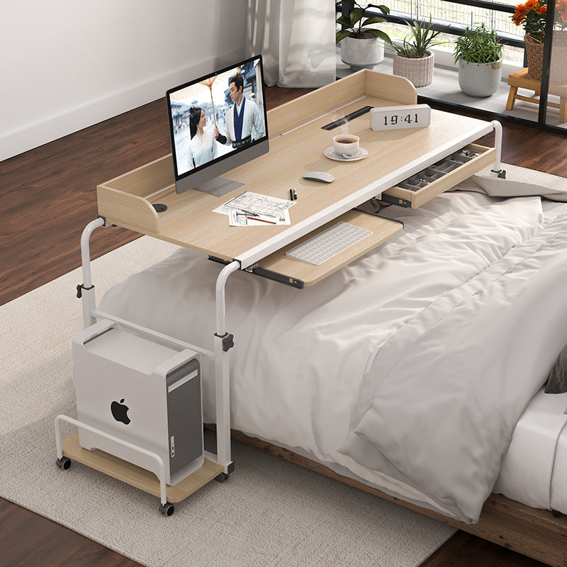 A white bed with a desk on wheels going across it that includes a laptop computer, keyboard, and mouse.